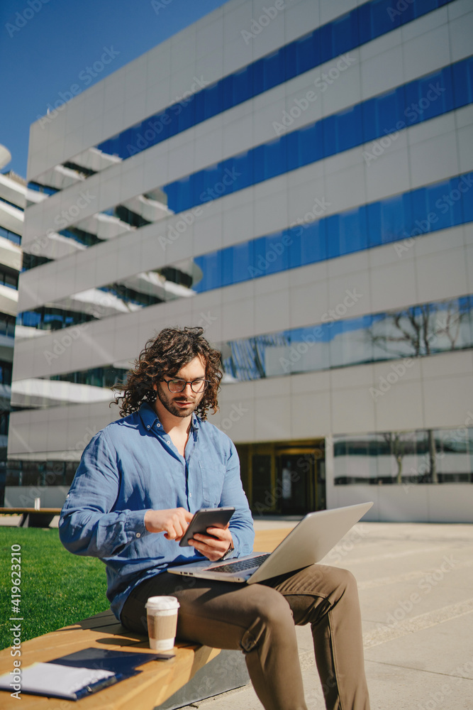 Handsome businessman with curly hair sitting on bench and using tablet Handsome businessman with curly hair sitting on bench and using tablet