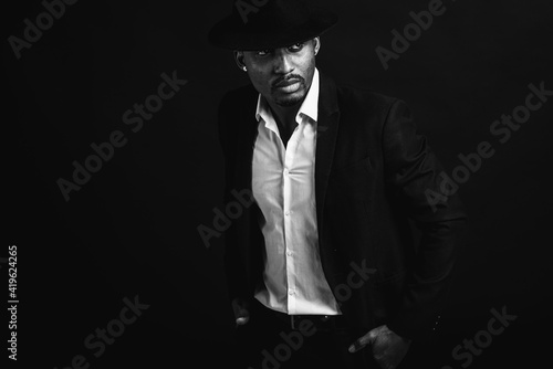 Portrait of attractive, cute, stylish professional African American businessman in stylish suit, white shirt and black hat, isolated on dark background. Low key. Selective focus