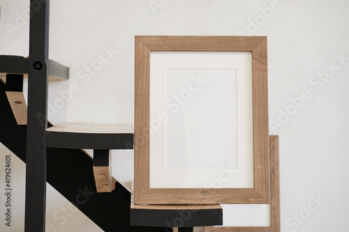Closeup of square empty wooden picture frame on wooden stairs. White wall background. Scandinavian interior, home design. Art concept. Artistic mockup scene. Front view