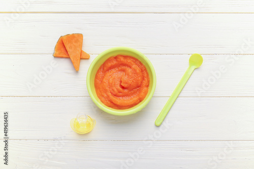 Bowl with pumpkin baby puree, spoon and pacifier on white wooden background
