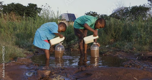 Water crisis. Close-up view of two young black African boys drinking water from a communal faucet and filing up plastic containers to take back home due to poor living conditions, no running water  