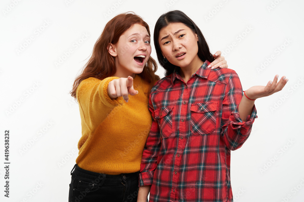 Two young women friends. Girl showing to her friend something, but another one can not see. Wearing yellow sweater and checkered shirt. Watching at the camera, isolated over white background Two young women friends. Girl showing to her friend something, but another one can not see. Wearing yellow sweater and checkered shirt. Watching at the camera, isolated over white background