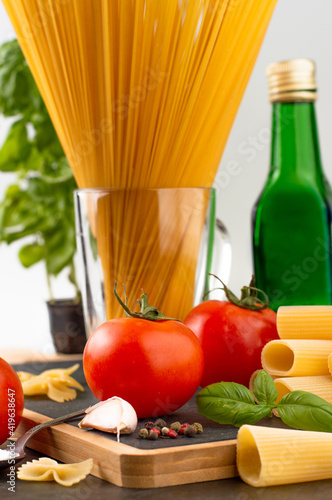 Blurred image in the foreground of a tomato, sprigs of basil, garlic, in the background spaghetti, olive oil.