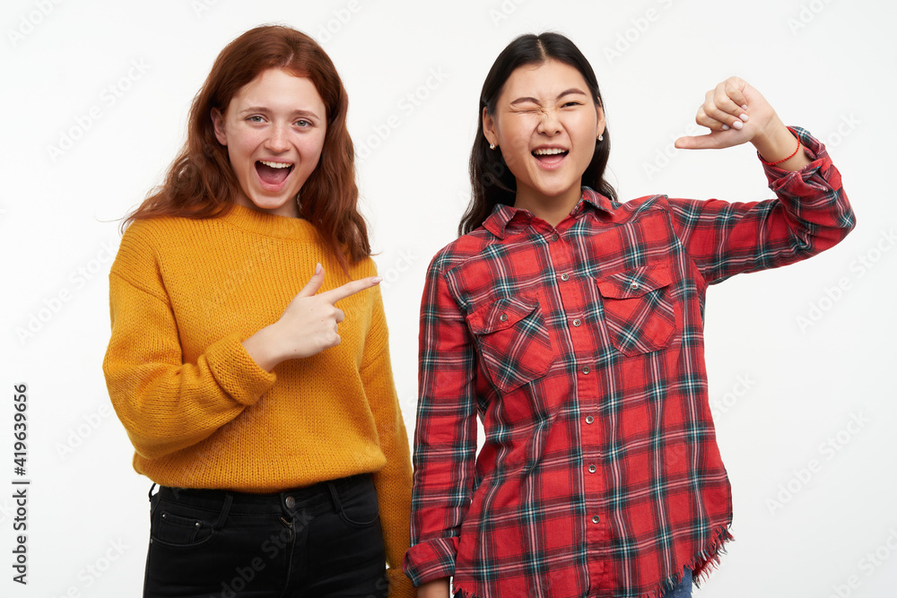 Portrait of cheerful asian and caucasian friends. Wearing casual outfit. Happy girl pointing on her friend who winks and pointing on her self. Watching at the camera, isolated over white background Portrait of cheerful asian and caucasian friends. Wearing casual outfit. Happy girl pointing on her friend who winks and pointing on her self. Watching at the camera, isolated over white background