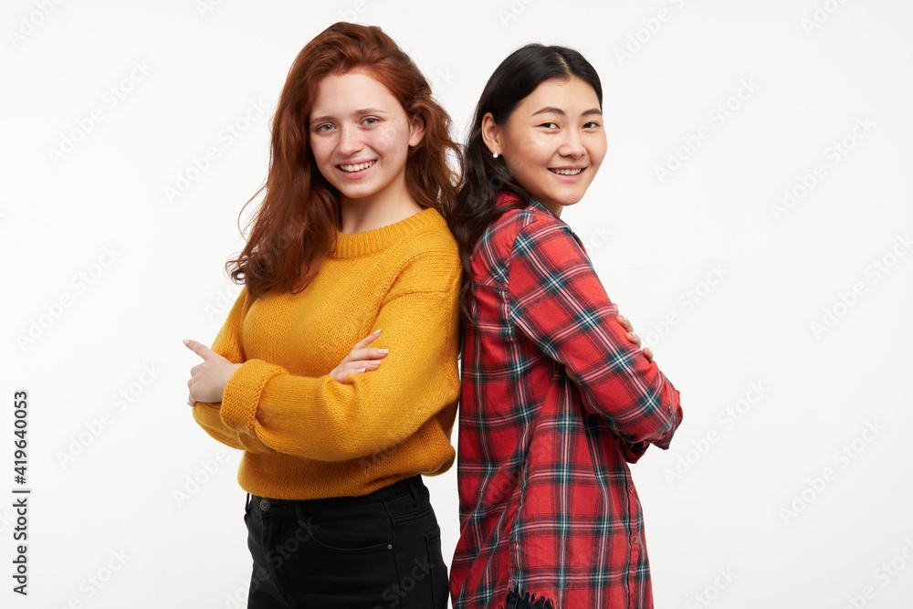 Two young women friends. Wearing yellow sweater and checkered shirt. Standing back to back with arms crossed. People and lifestyle concept. Watching at the camera, isolated over white background Two young women friends. Wearing yellow sweater and checkered shirt. Standing back to back with arms crossed. People and lifestyle concept. Watching at the camera, isolated over white background