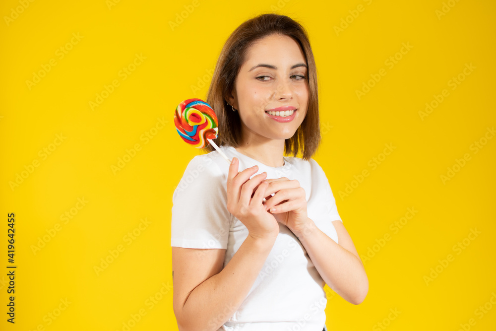 Beautiful young woman holding colorful lollipop candy in hand having fun over yellow background. Sweet food and happiness concept. Beautiful young woman holding colorful lollipop candy in hand having fun over yellow background. Sweet food and happiness concept.