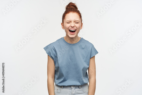 Funny woman, beautiful girl with ginger hair gathered in a bun. Wearing blue t-shirt and jeans. Laughing with closed eyes, hear hilarious joke. Emotion concept. Stand isolated over white background © timtimphoto