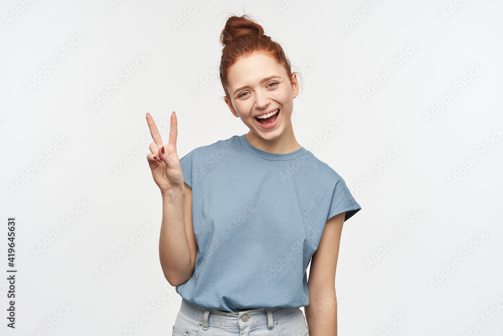 Portrait of cute, adult redhead girl with hair gathered in a bun. Wearing blue t-shirt and jeans. Showing peace sign, tilted her head and smile. Watching at the camera isolated over white background Portrait of cute, adult redhead girl with hair gathered in a bun. Wearing blue t-shirt and jeans. Showing peace sign, tilted her head and smile. Watching at the camera isolated over white background
