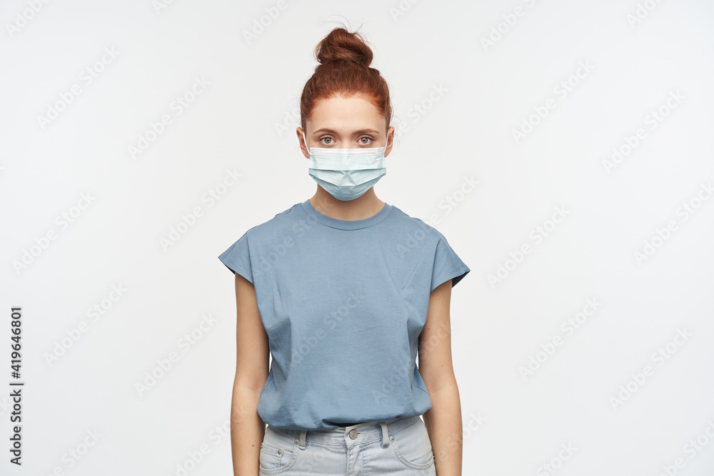 Teenage girl, serious looking woman with ginger hair gathered in a bun. Wearing blue t-shirt, jeans and protective face mask. Watching at the camera isolated over white background Teenage girl, serious looking woman with ginger hair gathered in a bun. Wearing blue t-shirt, jeans and protective face mask. Watching at the camera isolated over white background