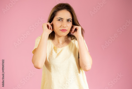 Young beautiful woman wearing casual t-shirt standing over isolated pink background covering ears with fingers with annoyed expression. Deaf concept.