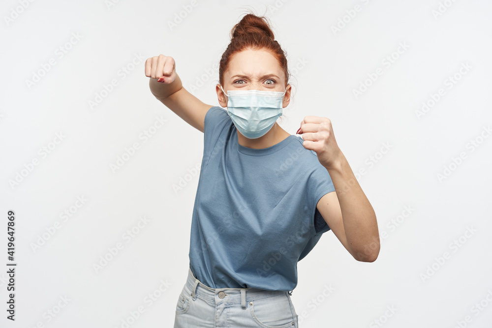 Aggressive looking woman with ginger hair gathered in a bun. Wearing blue t-shirt and protective face mask. Clench her fists and want to punch. Watching at the camera isolated over white background Aggressive looking woman with ginger hair gathered in a bun. Wearing blue t-shirt and protective face mask. Clench her fists and want to punch. Watching at the camera isolated over white background