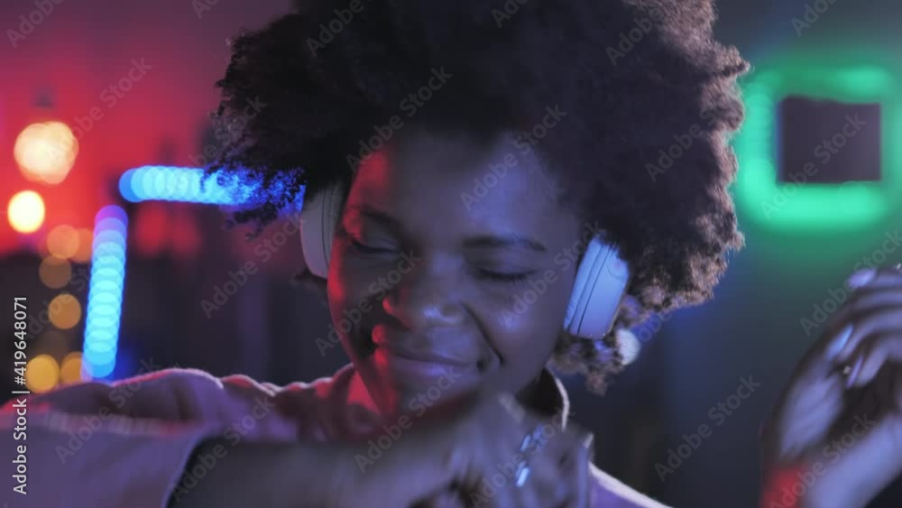 Black african girl in headphones dances in a dark room coloured led lights in the background,portrait of carefree curly afro american young woman listen to music, dancing with colorful neon lighting Black african girl in headphones dances in a dark room coloured led lights in the background,portrait of carefree curly afro american young woman listen to music, dancing with colorful neon lighting
