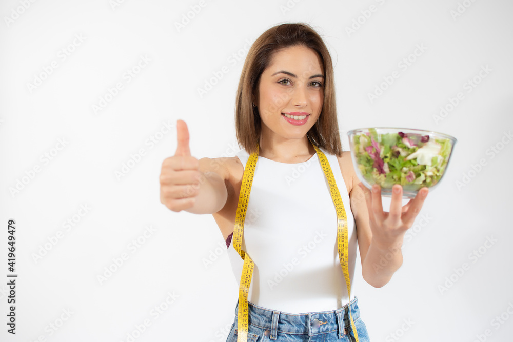 Young woman with measure tape showing a bowl of salad isolated over white background Young woman with measure tape showing a bowl of salad isolated over white background