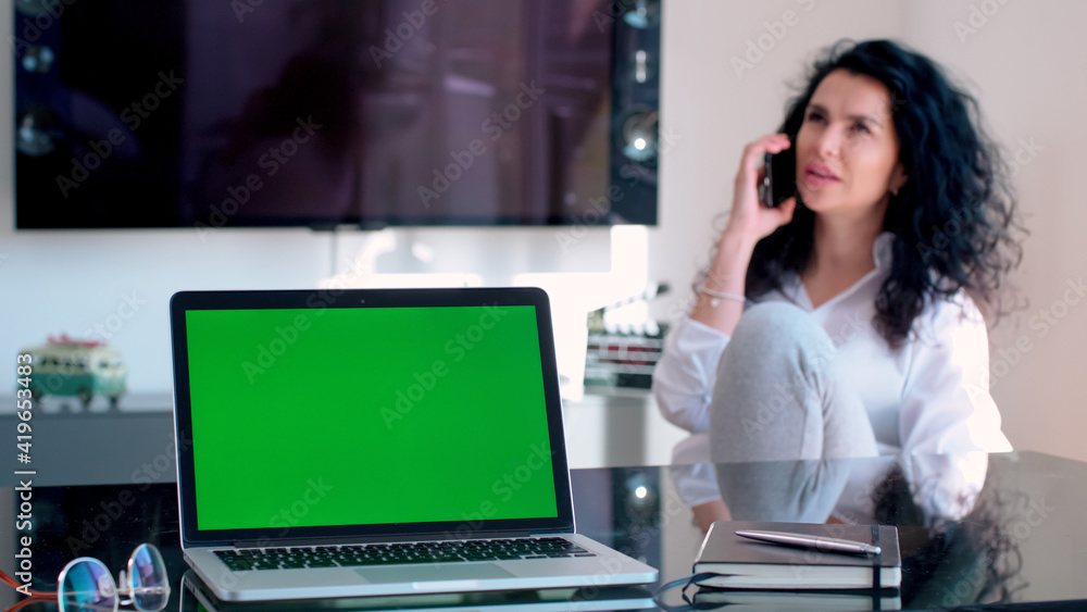 Beautiful multi ethnic young business woman on the phone in home office wear cozy jogging pants with green screen on laptop in foreground Beautiful multi ethnic young business woman on the phone in home office wear cozy jogging pants with green screen on laptop in foreground