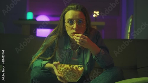 A shocked young girl with popcorn is watching a tv program or movie at night. Unexpected turn of events in a film. The amazed woman is popeyes surprised watching tv sitting on a couch at home. photo