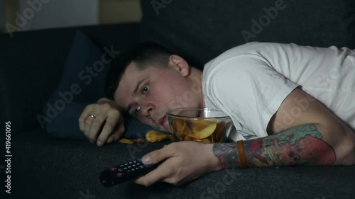 A man lying at coach and watching TV, eating fastfood