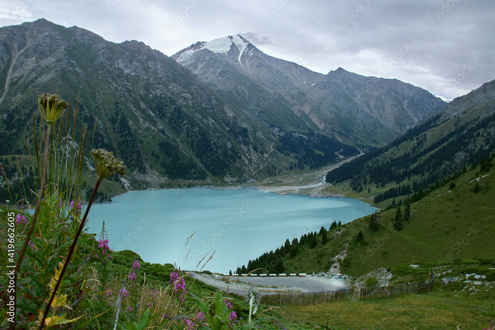 Alpine blue Big Almaty lake, summer day in the mountains, the sky in clouds, a lake surrounded by mountains, trees, grass and flowers grow on the mountainsides, a road passes near the lake