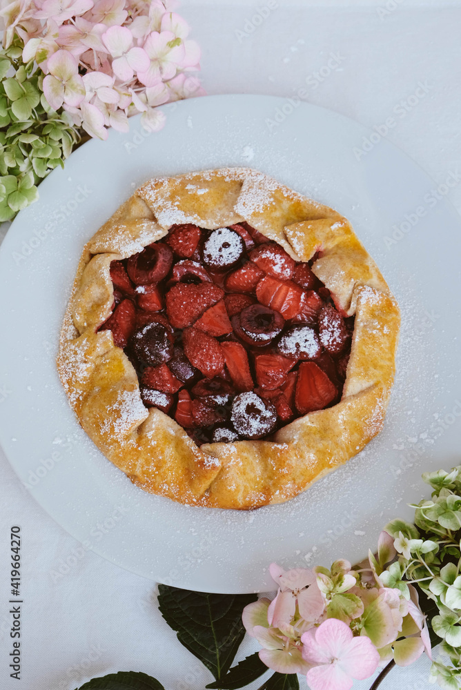 Delicious freshly baked vegan strawberry and cherry galette on white background with hydrangea flowers, top view. Sweet food, summer dessert.