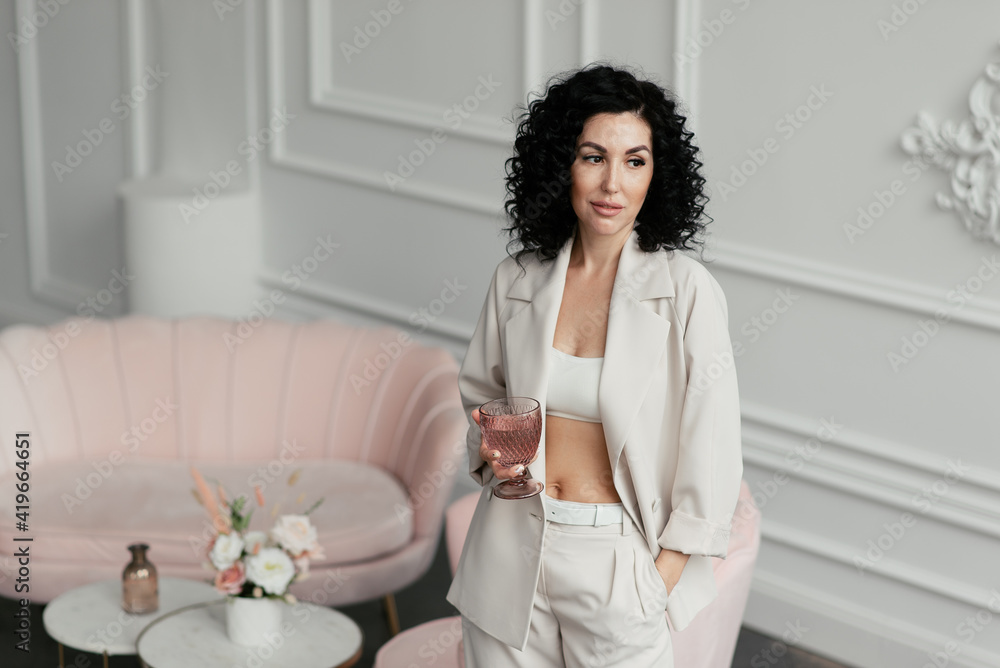 A woman with curly hair in a light suit with a naked belly holds a glass in her hands
