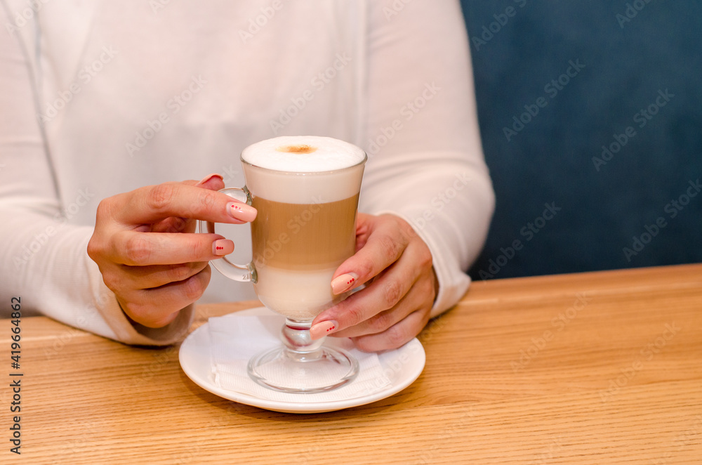 Young woman in a cafe drinks a latte, holds a transparent glass with her hand on a white saucer. Waits for the coffee to cool. With copyspace Young woman in a cafe drinks a latte, holds a transparent glass with her hand on a white saucer. Waits for the coffee to cool. With copyspace