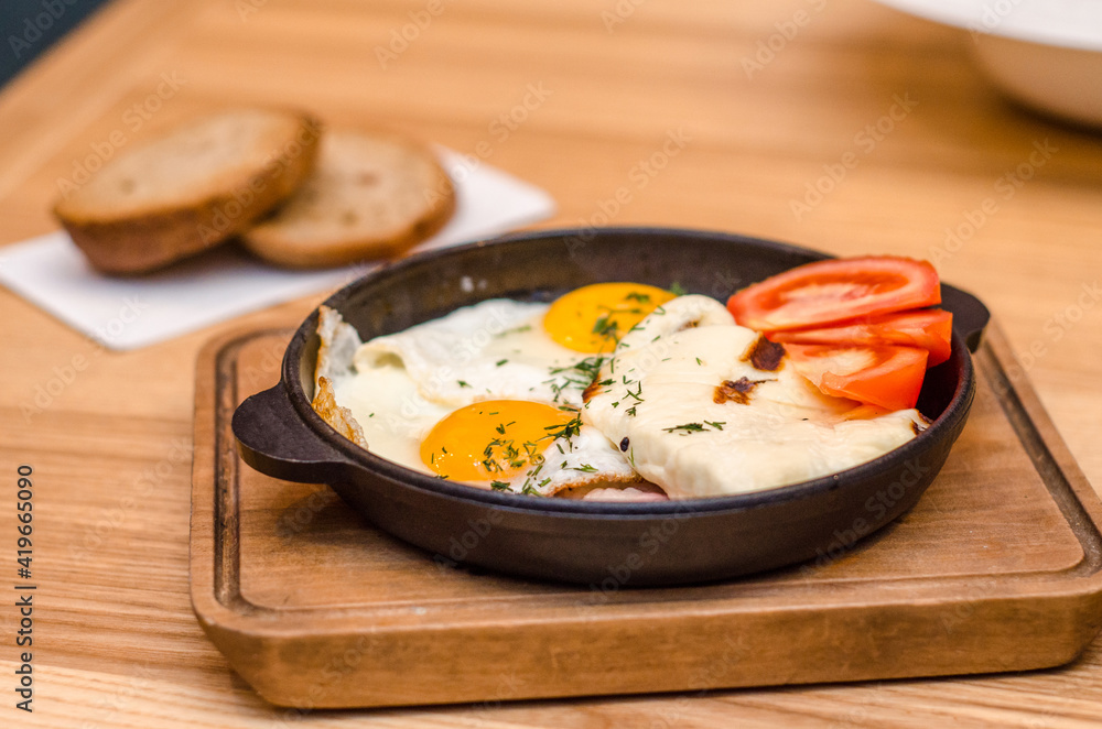 Hearty breakfast is served in a black frying pan and wooden board, fried eggs with bacon and tomatoes, two pieces of bread are on a wooden table Hearty breakfast is served in a black frying pan and wooden board, fried eggs with bacon and tomatoes, two pieces of bread are on a wooden table