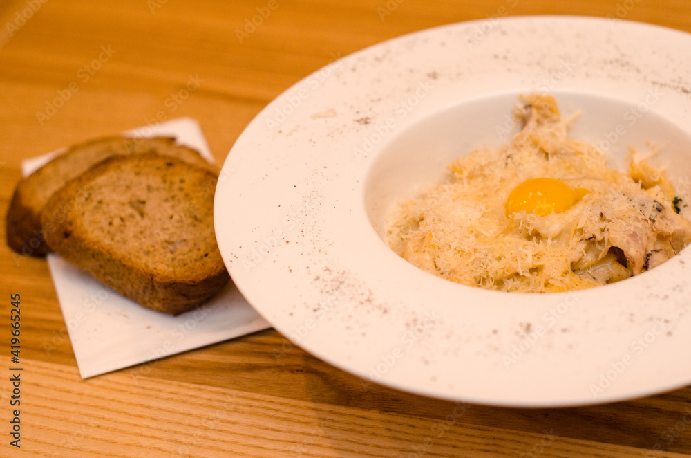 Appetizing carbonara pasta with egg yolk on a white plate in a restaurant, two pieces of bread lie on a wooden table Appetizing carbonara pasta with egg yolk on a white plate in a restaurant, two pieces of bread lie on a wooden table
