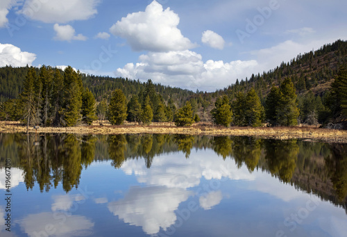 Original landscape photograph of a glassy lake reflecting the evergreen tree covered hills and white billowy clouds