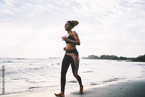 Happy young female in active clothes reaching running goals on cardio workout in morning  slim dark skinned energetic sportive woman training athletic strength outdoors jogging around seashore