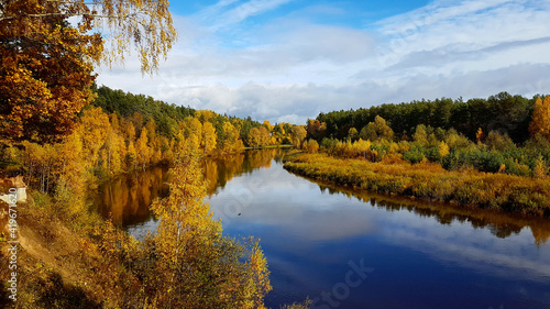 Orange autumn landscape overlooking a calm river with a reflection of blue sky surrounded by forest. Trees have yellow leaves.
