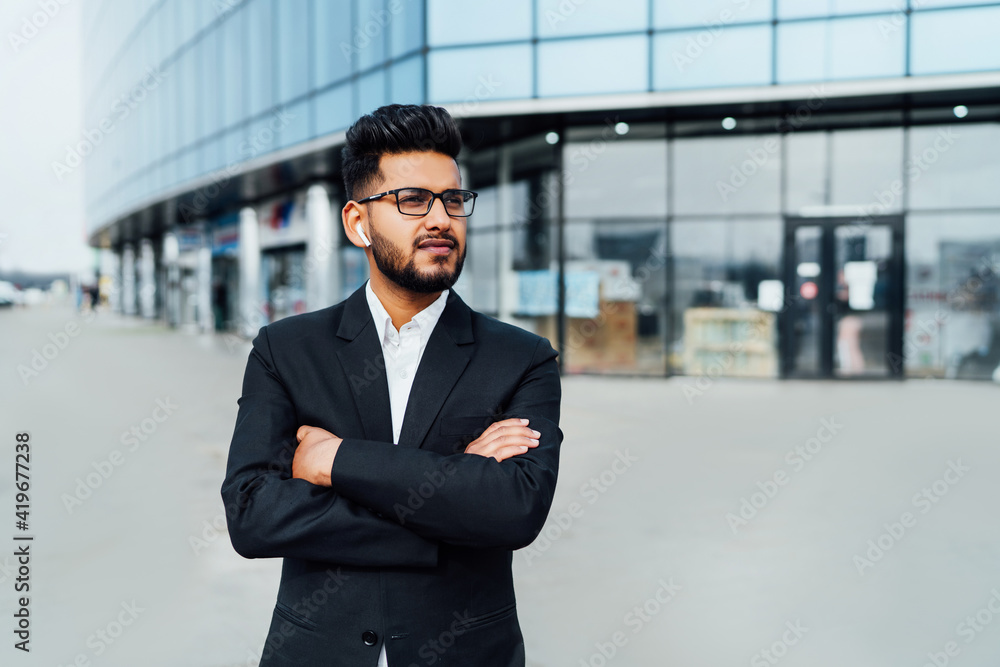 Fototapeta premium A modern office worker, an Indian man, a bank employee in a black suit, behind a large wooden building