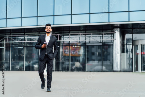 A smiling bearded Indian man walks to an important meeting, behind him a modern building, he smiles and looks at the camera