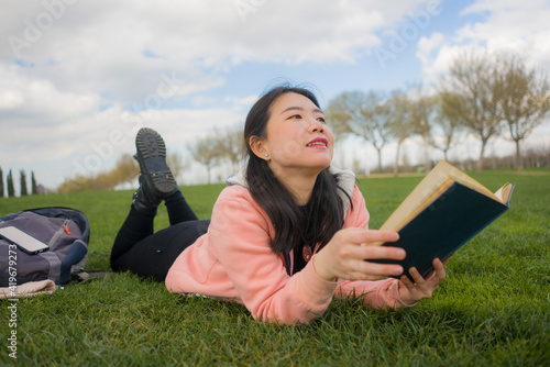 young Asian woman enjoying novel on grass - lifestyle portrait of young happy and pretty Japanese girl reading a book at beautiful city park in reading and studying concept © TheVisualsYouNeed