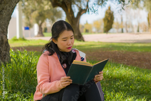 young Asian woman enjoying novel on grass - lifestyle portrait of young happy and pretty Japanese girl reading a book at beautiful city park in reading and studying concept