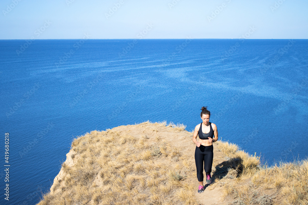 Tired young athletic woman running hard at mountain landscape nature path near blue ocean in summer sunny day. Wearing black sportswear. Weight loss cardio goal achievement challenge. Copy space