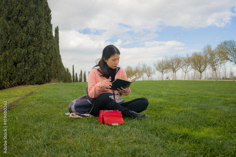 Fototapeta premium young Asian woman enjoying novel on grass - lifestyle portrait of young happy and pretty Chinese girl reading a book at beautiful city park in reading and studying concept