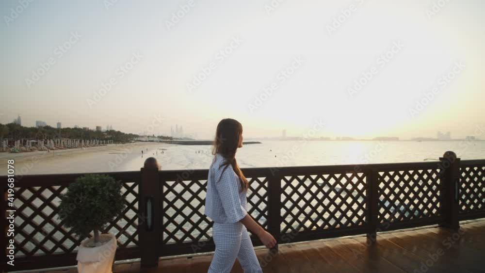 Woman walking on ocean pier in Dubai during sunset. Side view of woman looking at Burj Al Arab hotel and sand beach from a wooden pier Woman walking on ocean pier in Dubai during sunset. Side view of woman looking at Burj Al Arab hotel and sand beach from a wooden pier
