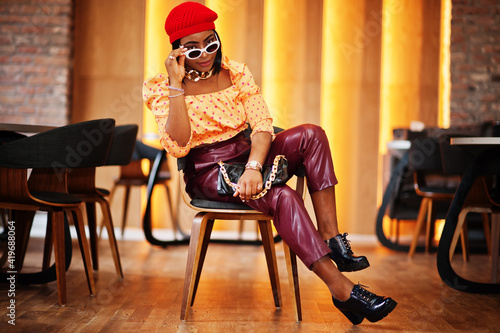 Elegant afro american woman in red french beret, big gold neck chain, sunglasses, polka dot blouse and leather pants pose indoor. © AS Photo Family