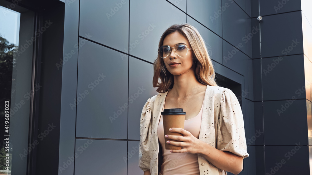 Blonde woman in sunglasses holding coffee to go while walking near building Blonde woman in sunglasses holding coffee to go while walking near building