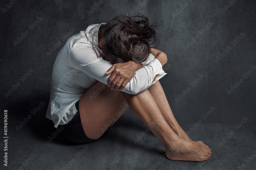 Depression or domestic violence concept: Sad lonely young woman crying while sitting on the floor in a dark room with an attitude of sadness and boredom with her legs together and hugged. Depression or domestic violence concept: Sad lonely young woman crying while sitting on the floor in a dark room with an attitude of sadness and boredom with her legs together and hugged.