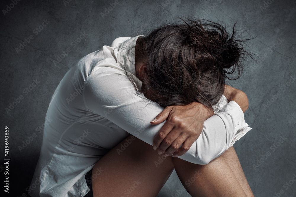 Depression or domestic violence concept: Sad lonely young woman crying while sitting on the floor in a dark room with an attitude of sadness and boredom with her legs together and hugged. Depression or domestic violence concept: Sad lonely young woman crying while sitting on the floor in a dark room with an attitude of sadness and boredom with her legs together and hugged.