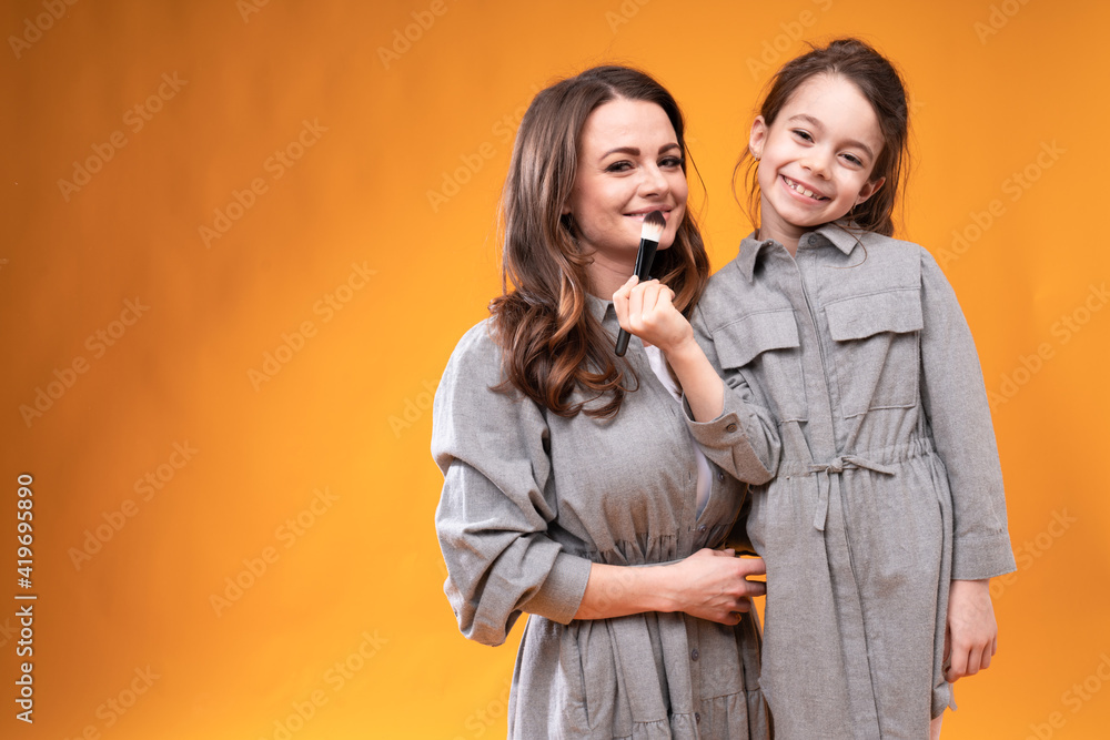 Daughter in a gray dress smiles and makes makeup to a young mother using a brush while standing on a bright background Daughter in a gray dress smiles and makes makeup to a young mother using a brush while standing on a bright background