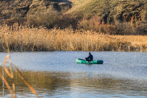 A man in an old inflatable boat is fishing in the lake with a fishing rod, but against the background of yellow reeds