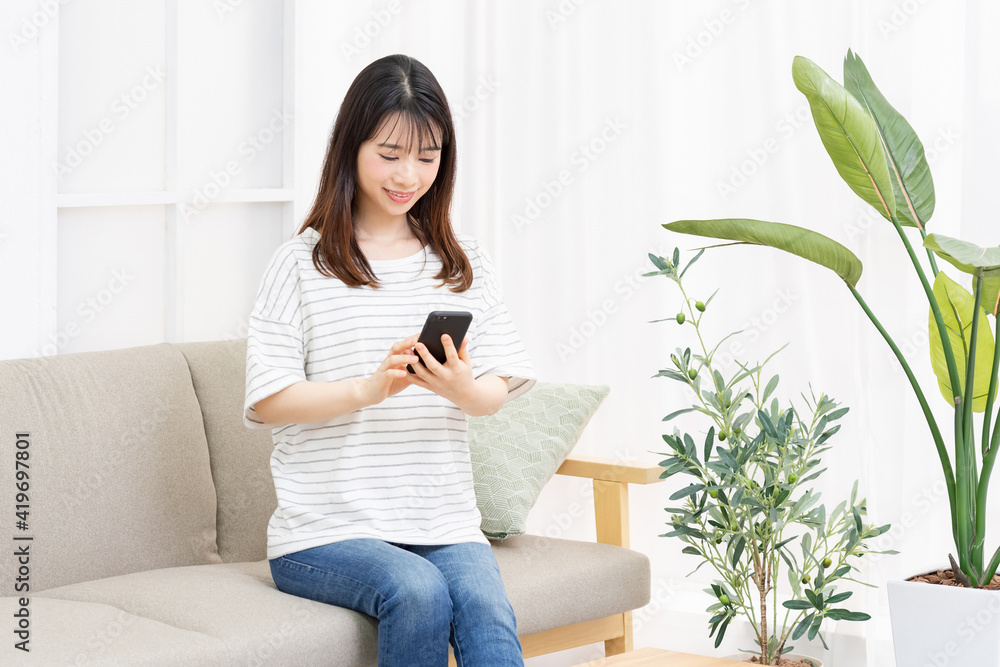 部屋のソファーに座ってスマホを操作する若い女性 部屋のソファーに座ってスマホを操作する若い女性