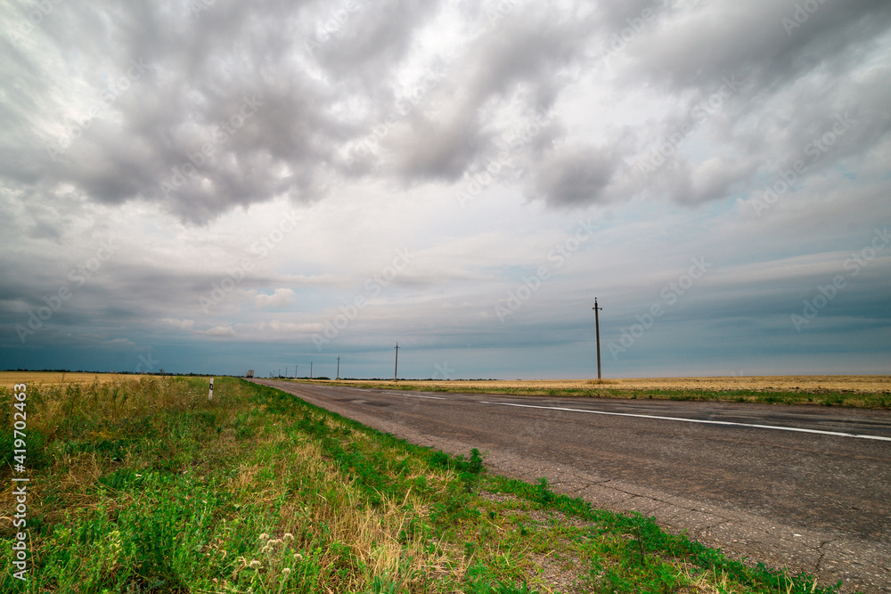 an asphalt road among the fields over which thunderclouds hang