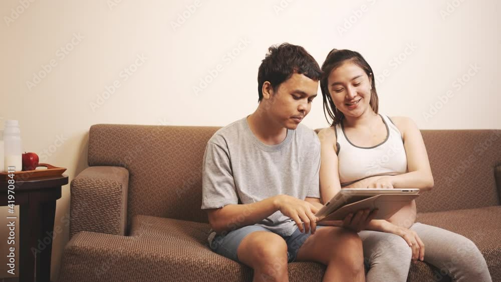 Happy Pregnant woman and husband shopping online baby clothes with tablet at home sitting on sofa internet technology, New family baby born concept. Happy Pregnant woman and husband shopping online baby clothes with tablet at home sitting on sofa internet technology, New family baby born concept.