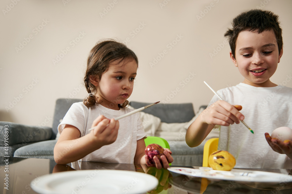 Two adorable children, a schoolboy and a schoolgirl with braids, are sitting side by side decorating Easter eggs. Two adorable children, a schoolboy and a schoolgirl with braids, are sitting side by side decorating Easter eggs.