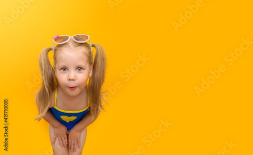 Little funny child in a blue swimsuit grimaces on a yellow background. A cute girl with ponytails on her head and sunglasses looks into the frame in surprise. Horizontal format. Place for text