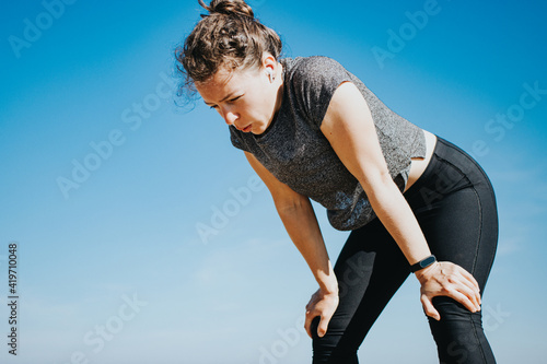 Tired young athletic woman runner taking a rest, doing break, breathing hard after running hard outdoors, standing on blue sky background. Weight loss cardio goal achievement challenge. Copy space