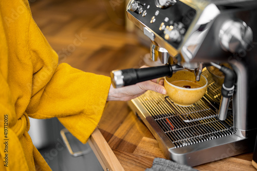 Woman in yellow bathrobe making espresso drink on a professional coffee machine at home, close-up. The process of making coffee on a carob machine.