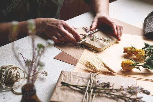 Woman makes zero waste, plastic free, trendy hand made gift package with craft recycled paper and dried flowers on the table with linen tablecloth. Natural aesthetic.